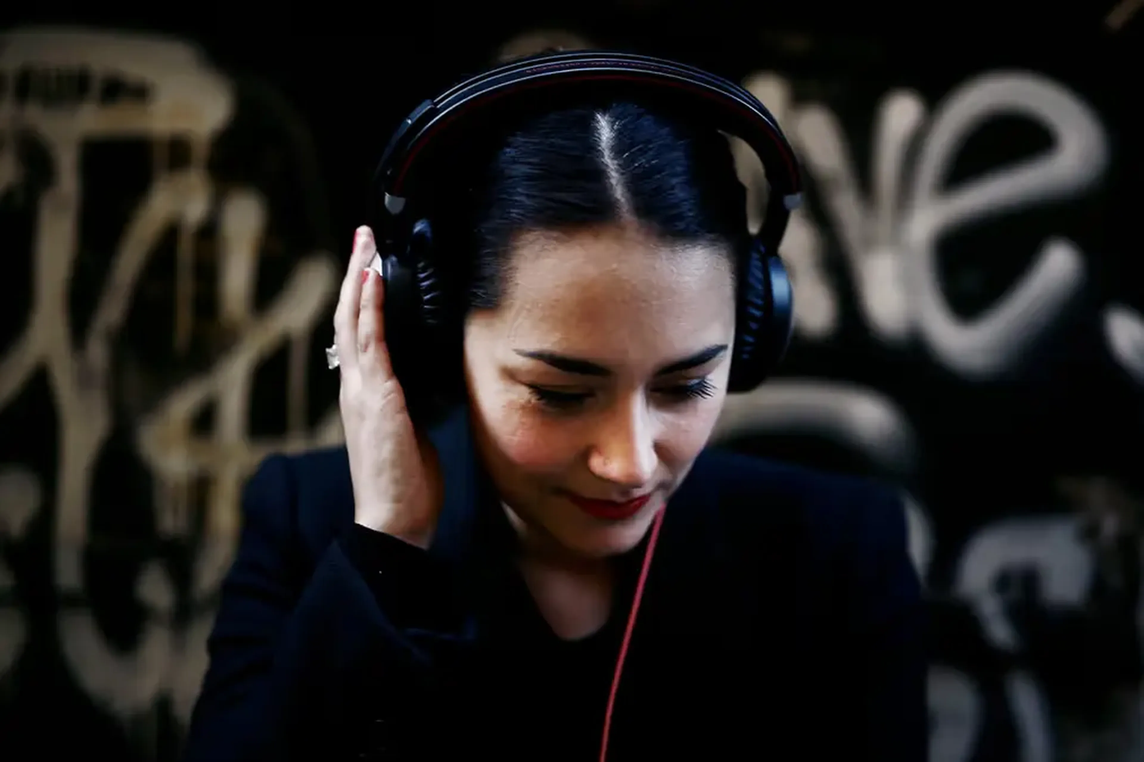 Woman with headphones, eyes closed, wearing a black jacket, stands against a graffiti-covered wall.