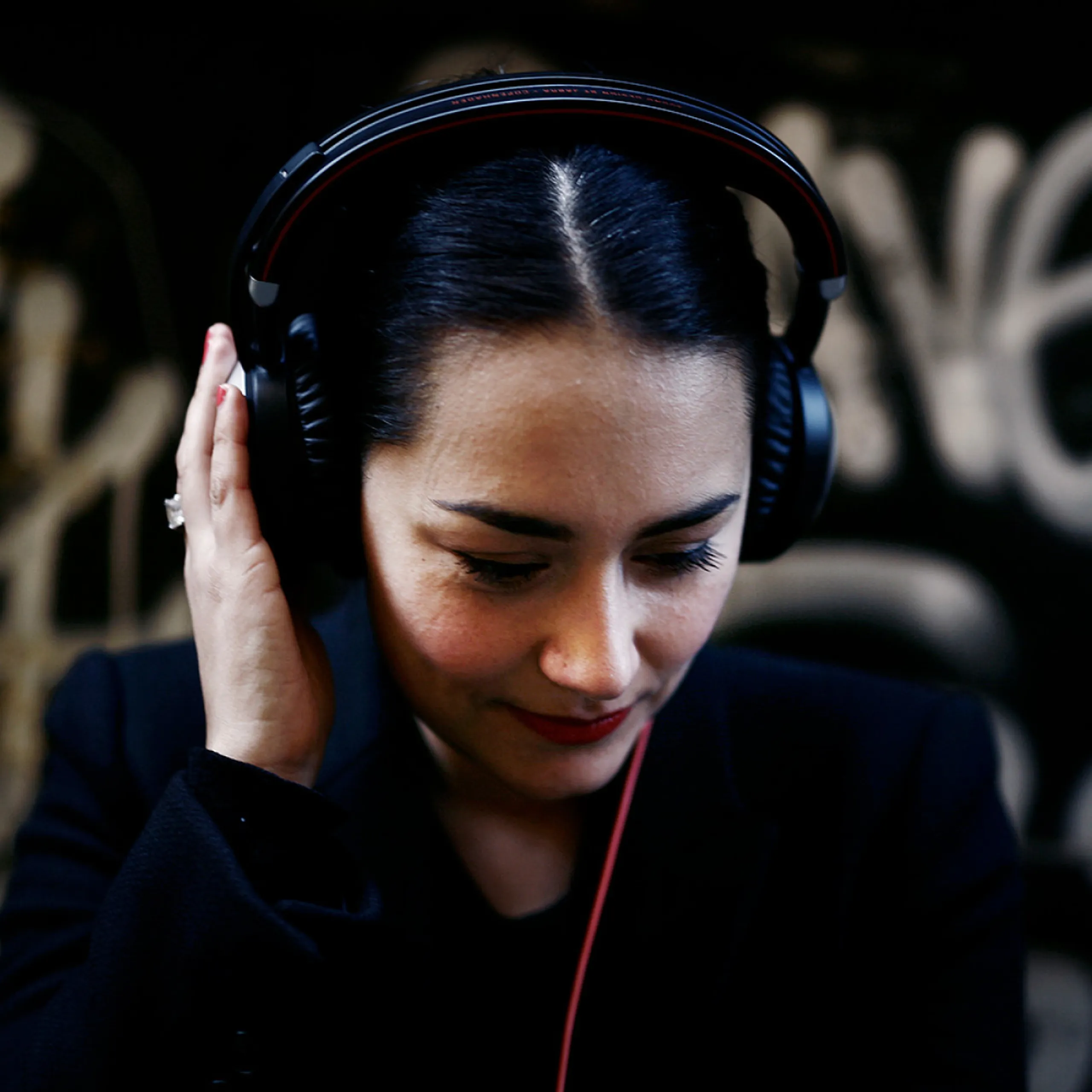 Woman with headphones listening, against a dark graffiti wall.
