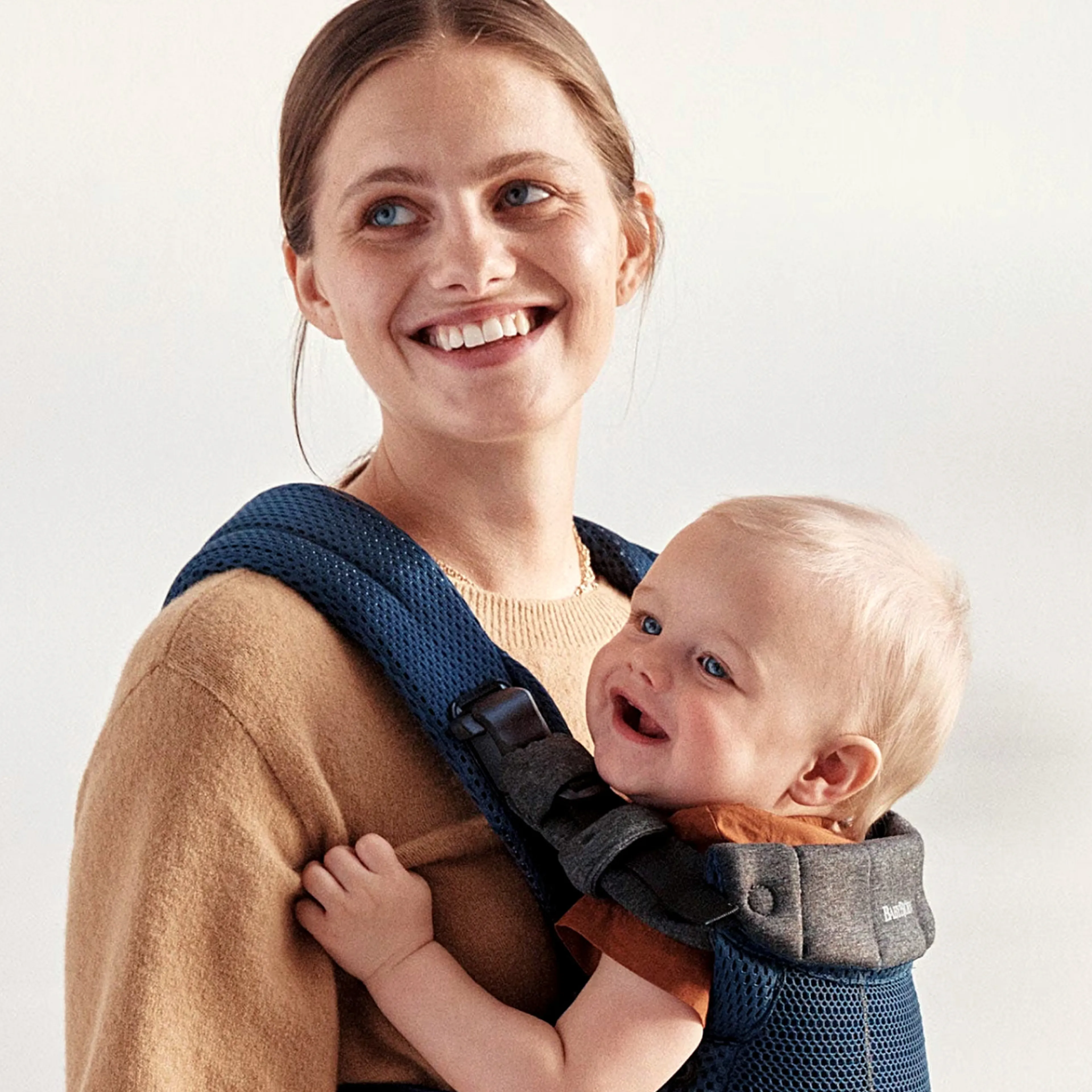 Smiling woman carrying a happy baby in a blue baby carrier, both looking content, against a neutral background.