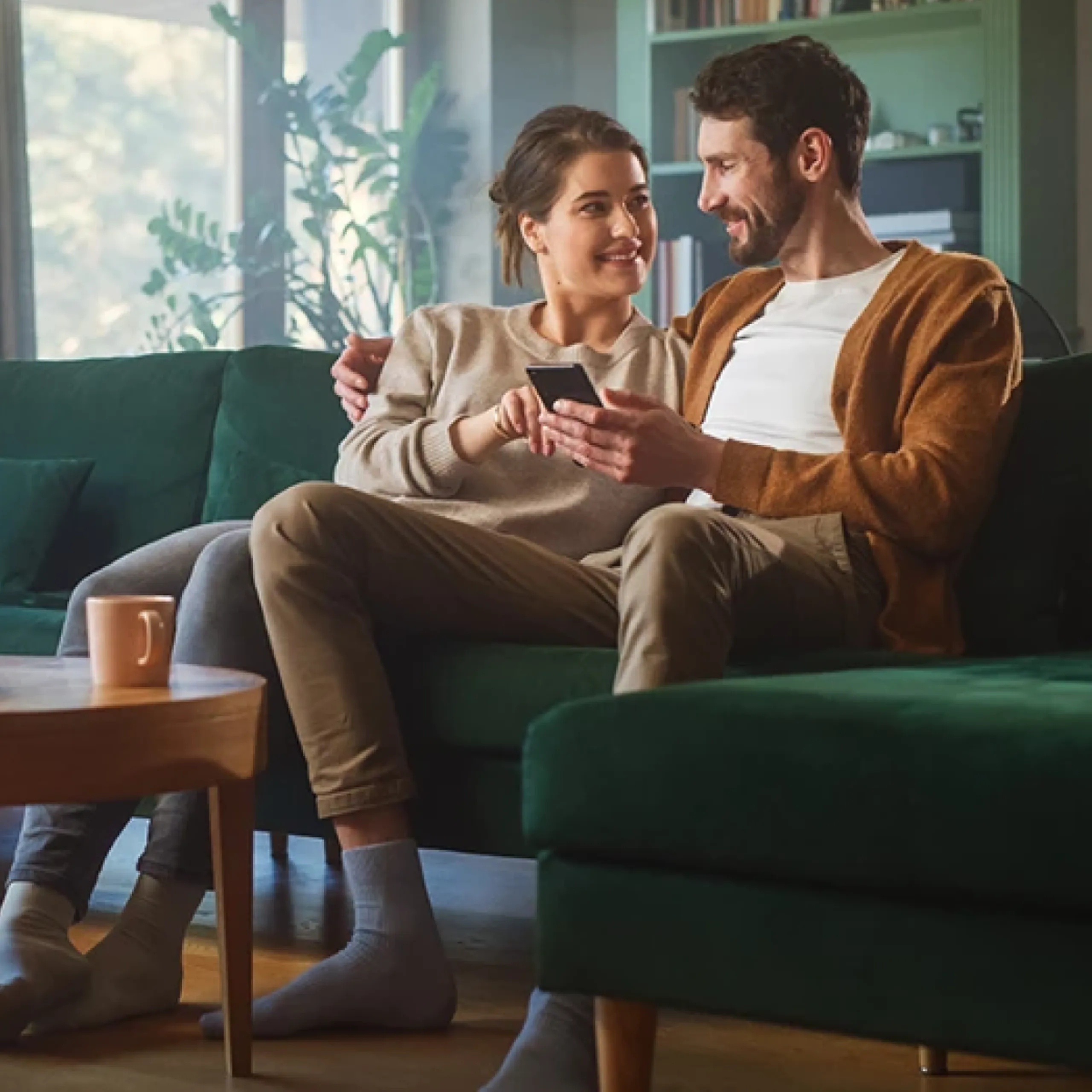 Couple smiling at each other on a green sofa while looking at a phone.