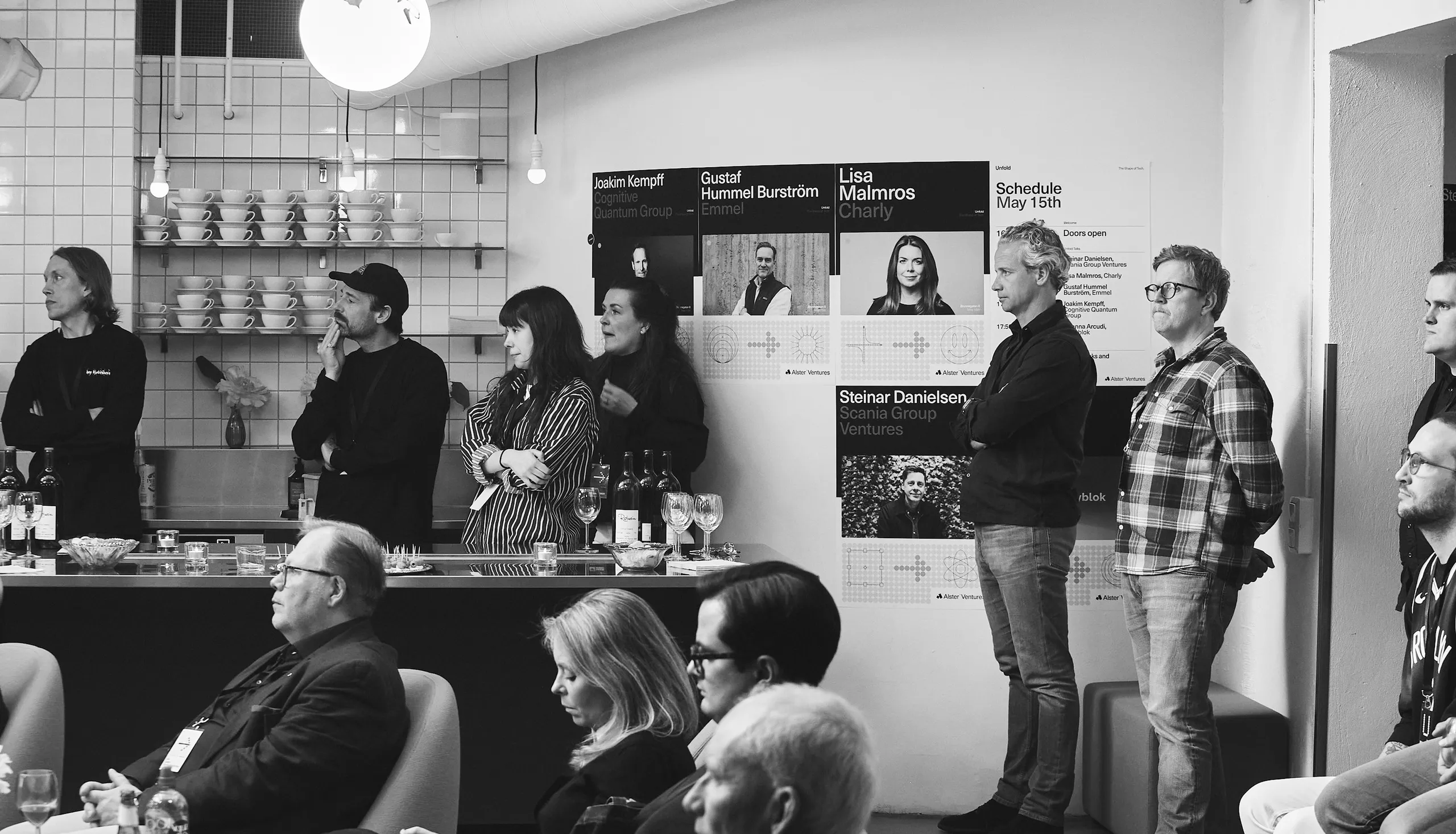 Guests at an Alster Ventures event, standing and sitting in a room with speaker posters on the wall.